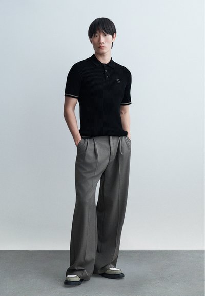 Άνδρας που φοράει μαύρο κοντομάνικο ριμπ μπλουζάκι πόλο και φαρδιές γκρι πλισέ παντελόνες, στέκεται με τα χέρια στις τσέπες σε ένα απλό φόντο.