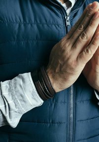 Brown leather bracelets are stacked on a wrist, varying in textures and thickness. A blue puffer vest and a striped shirt are visible.