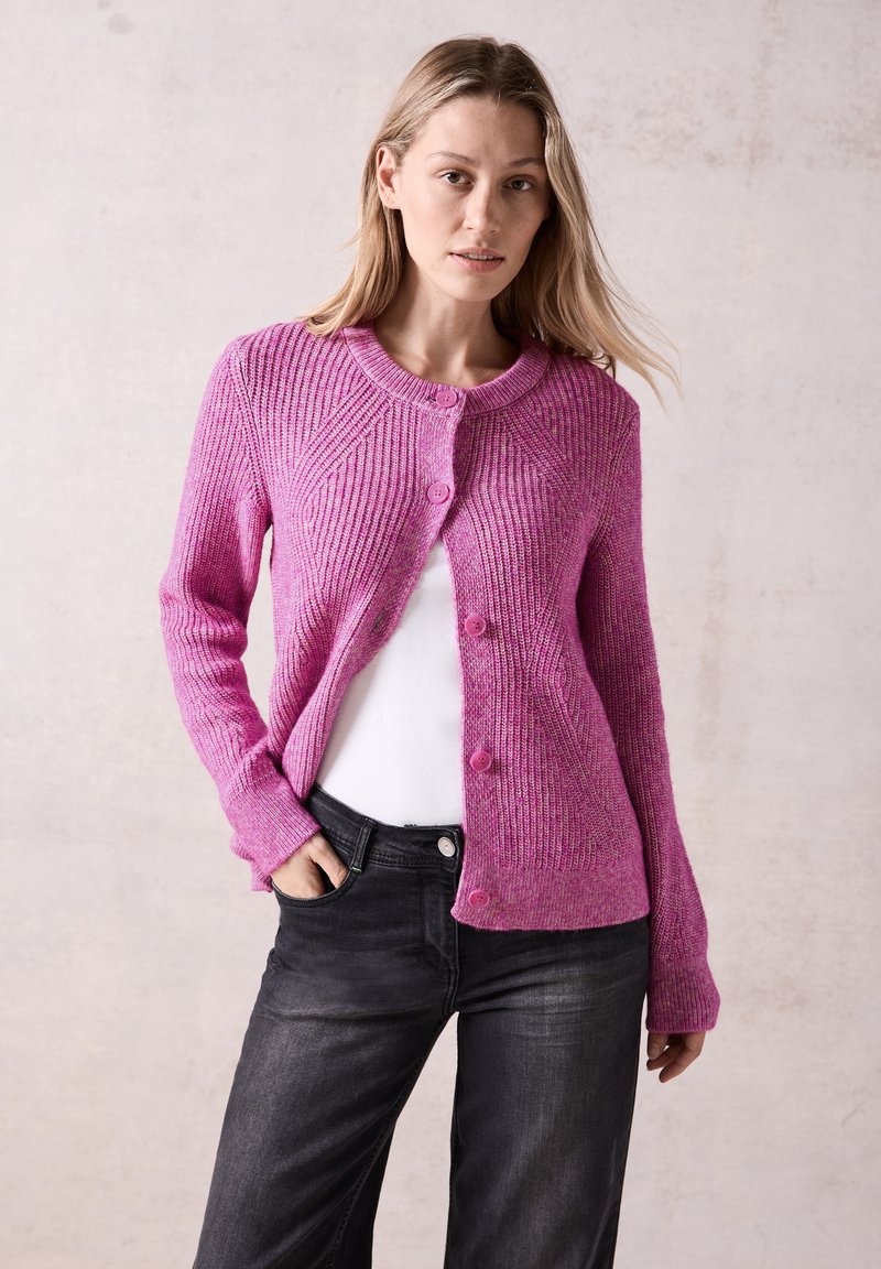 Růžový žebrovaný pletený cardigan s kulatým výstřihem, uzávěrem na knoflíky a dlouhými rukávy, ve spojení s bílou košilí a tmavými džíny.