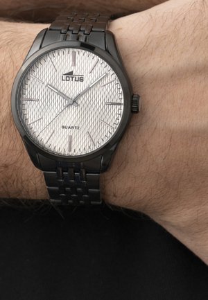 Crni metalni ručni sat s teksturiranim bijelim brojčanikom, srebrnim oznakama sati i imenom marke "Lotus", nošen na lijevom zapešću osobe.