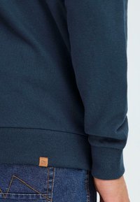 Tums zilā sporta krekls izgatavots no mīksta kokvilnas, ar ribotām aprocēm un apakšmalu, ar mazu brūnu logotipa etiketi tuvu apakšam.