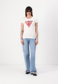 Witte aansluitende t-shirt met een bloemmotief en grafisch driehoekig ontwerp, gecombineerd met lichtblauwe flared jeans en zwarte flats; model draagt een zonnebril.