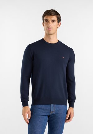 Uomo che indossa un maglione blu navy a maniche lunghe e jeans blu in piedi davanti a uno sfondo bianco semplice.