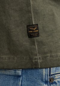 Chemise en coton vert olive avec une étiquette noire et dorée portant l'inscription "PME Legend", associée à un jean en denim bleu clair et un bouton en métal.
