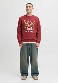 Rode sweatshirt met cartoon rendier ontwerp, met het uiterlijk van een rendier met gewei en versieringen, en de tekst "Laat het sneeuwen," gecombineerd met losse blauwe jeans en beige schoenen.