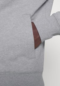 Sudadera gris con bolsillo tipo canguro, puños acanalados y textura suave. La abertura del bolsillo muestra un forro interior marrón.