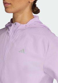 Veste zippée lilas avec capuche, texture lisse et petit logo gris sur la poitrine. Design léger, idéal pour le superposition.