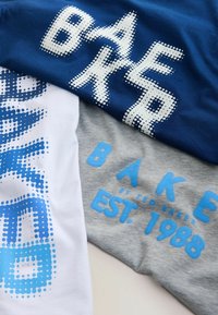 Trois t-shirts empilés : bleu marine avec texte ponctué blanc et turquoise, blanc avec des pois bleus, et gris avec des détails bleus "BAKE" et "EST. 1988".