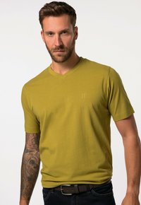 Camiseta de manga corta y cuello en V en algodón color verde oliva con un sutil detalle de logo. Textura suave, diseño ajustado, combinada con unos jeans oscuros y un cinturón.