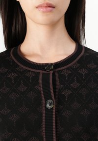 Cardigan en tricot noir avec un motif géométrique discret en marron foncé. Col côtelé et fermeture à boutons avec un détail de bouton circulaire brillant.