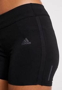 Pantalones cortos deportivos negros hechos de tela elástica, con un logo de Adidas, costuras planas y rayas laterales en contraste para mayor visibilidad.