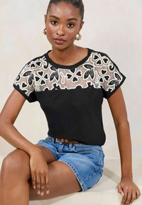 Top negro con un yoke de encaje que presenta patrones florales; mangas cortas; combinado con pantalones cortos de mezclilla. Tejido texturizado y diseño ajustado.