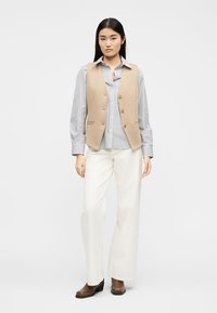 Γυναίκα με μπεζ γιλέκο με κουμπιά πάνω από ριγέ πουκάμισο, άσπρο παντελόνι με φαρδύ πόδι και καφέ μπότες με τακούνι, στέκεται μπροστά σε απλό φόντο.