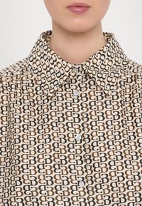 Femme portant une chemise beige avec un motif répétitif double-B marron et noir et un col pointu, boutonnée à l'avant.