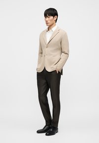 Άνδρας που στέκεται με τα χέρια στις τσέπες φορώντας μπεζ σακάκι, λευκό πουκάμισο, σκούρο παντελόνι και μαύρα παπούτσια, σε απλό φωτεινό φόντο.