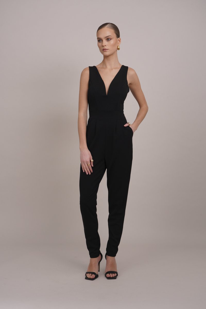 Mono negro con un profundo escote en V, cuerpo ajustado, piernas en tapered y bolsillos laterales. Combinado con tacones negros de tiras y accesorios dorados.