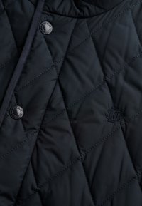 Quiltet marineblå pufferjakke stof med diamantmønster, der har knaplukning og broderet logo. Glat, let tekstur.