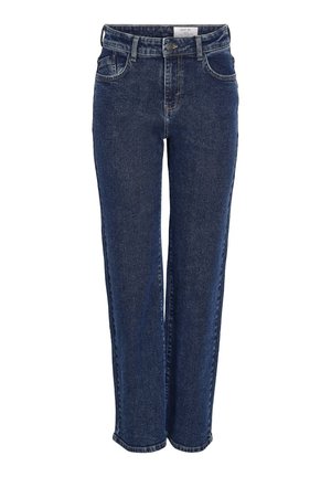 Femme portant un haut blanc sans manches, un jean droit bleu foncé et des baskets argentées, debout devant un fond uni.