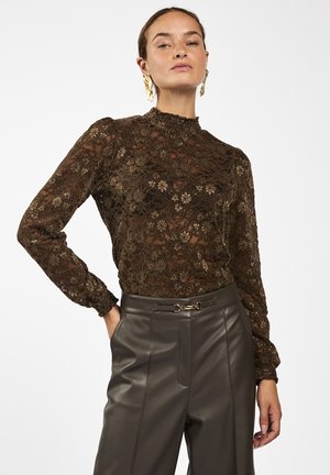 Vrouw die een bruine bloemrijke kanten blouse met lange mouwen draagt en een hooggetailleerde donkere leren broek met een decoratieve riem.