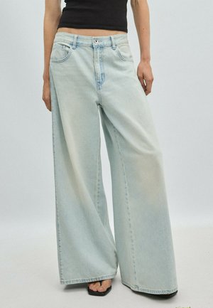 Person iført lyseblå jeans med brede ben og høj talje samt sorte åbne sandaler, stående mod en ensfarvet lys baggrund.