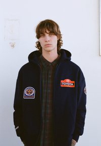 Tmavomodrá mikina s kapucňou s vyšívanými nášivkami Toyota, vrátane nápisu "Land Cruiser" a červeného loga Toyota, cez košeľu s károvaným vzorom a gombíkmi.