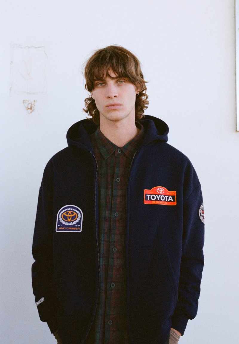 Tmavomodrá mikina s kapucňou s vyšívanými nášivkami Toyota, vrátane nápisu "Land Cruiser" a červeného loga Toyota, cez košeľu s károvaným vzorom a gombíkmi.