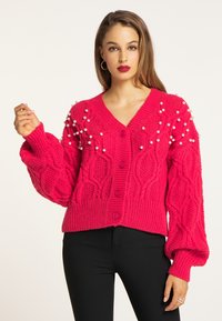 Cardigan tricotat roz intens, cu un model texturat de cabluri și accente de perle, cu decolteu în V și închidere cu nasturi. Mâneci lungi și voluminioase.