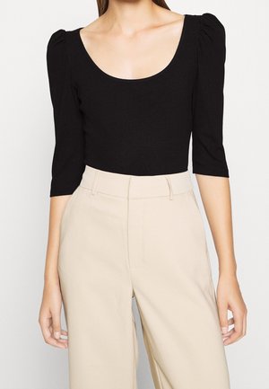 Femme portant un haut noir ajusté avec des épaules bouffantes et des manches trois-quarts, associé à un pantalon beige taille haute à jambes larges.