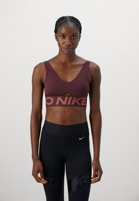 Egy nő áll, szemben nézve, bordó Nike sportmelltartóban és fekete Nike Pro leggingsben, egy sima háttér előtt.