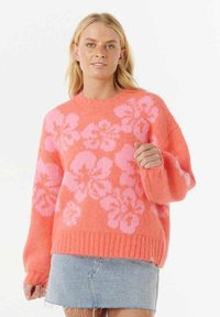 Frau, die einen leuchtend orangefarbenen Pullover mit pinken Blumenmustern und einen hellblauen Jeansrock trägt, steht vor einem einfarbigen weißen Hintergrund.