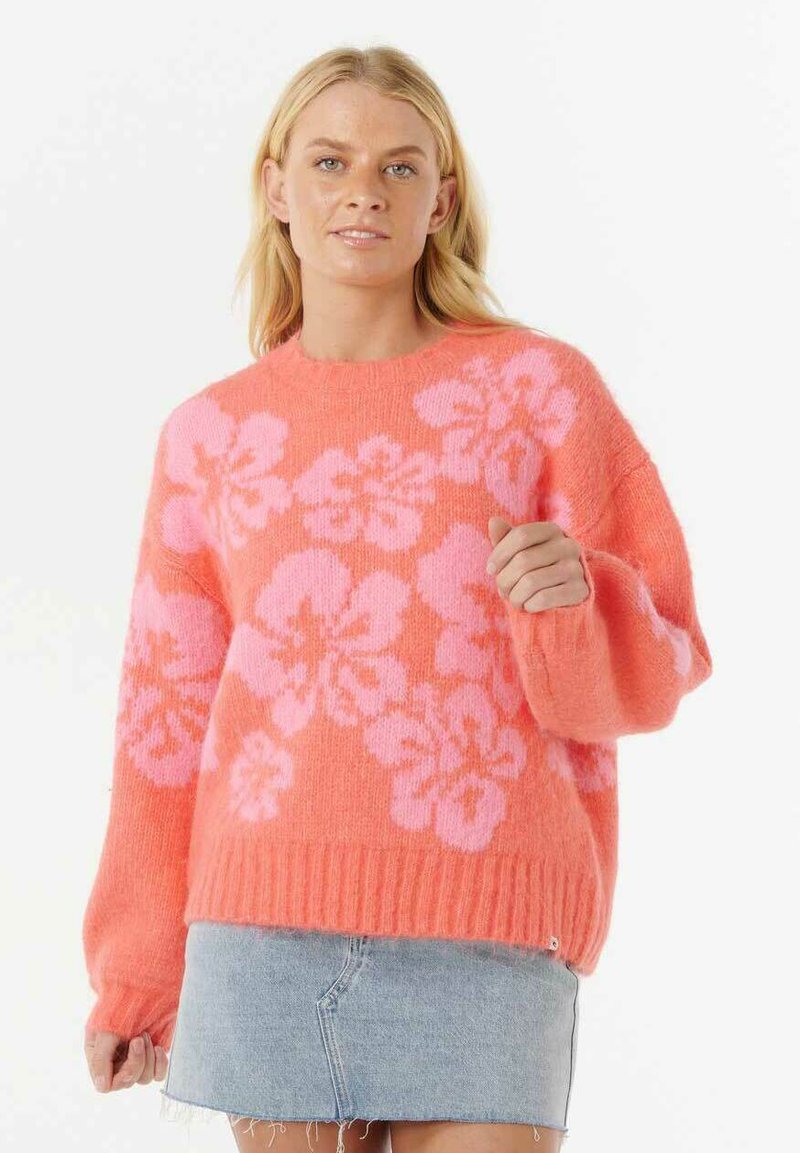 Femme portant un pull orange vif avec des motifs floraux roses et une jupe en denim bleu clair, se tenant contre un fond blanc uni.
