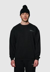 Svart bomullströja med sänkt axel och ribbade ärmslut, med en liten vit logotyp på den övre vänstra bröstområdet. Tillsammans med en svart mössa.