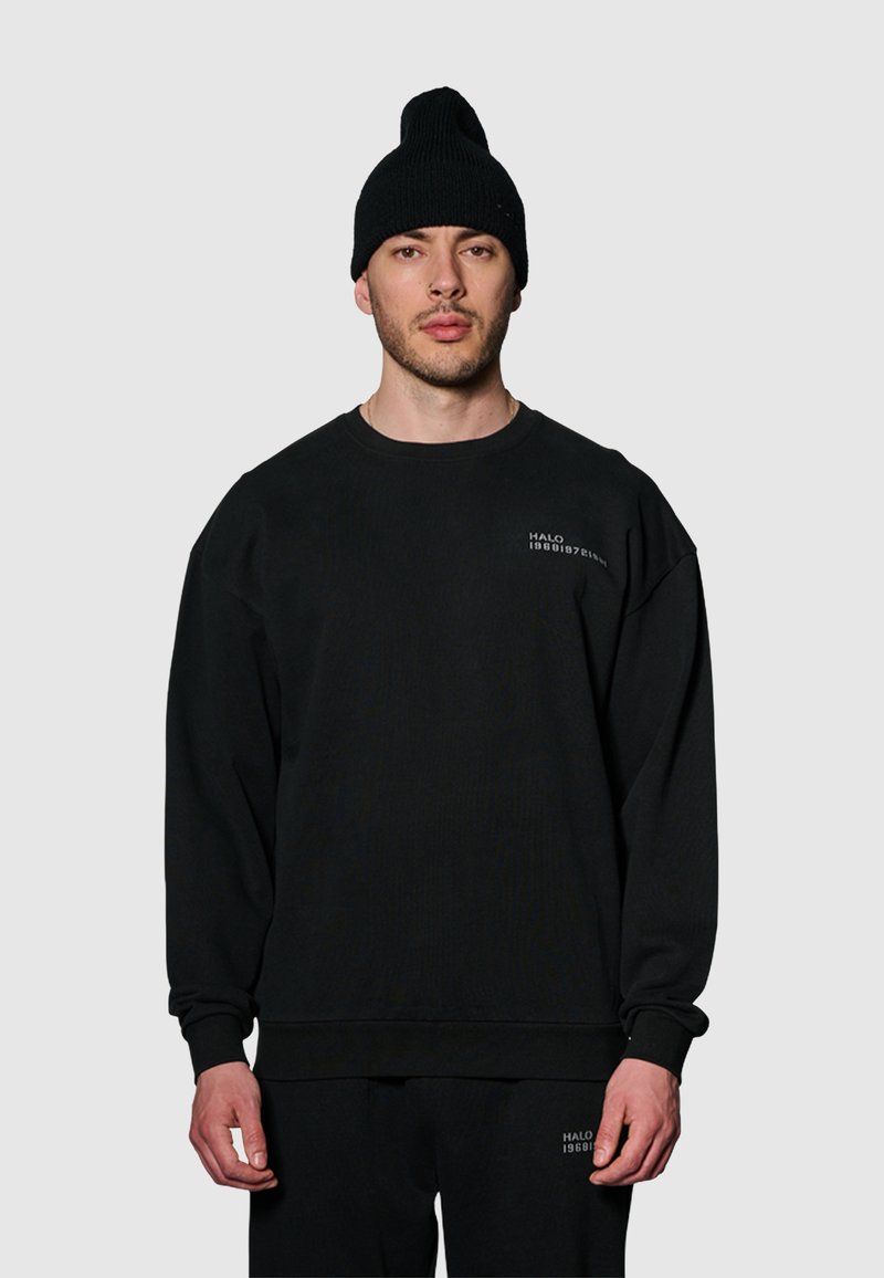 Svart bomullströja med sänkt axel och ribbade ärmslut, med en liten vit logotyp på den övre vänstra bröstområdet. Tillsammans med en svart mössa.