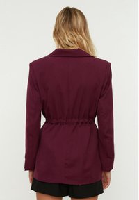 Bordový blazer s přiléhavým pasem, dlouhými rukávy a klopovým límcem. Hladká textura s nařaseným detailem na zádech, propracovaný design.