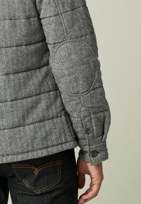 Grå herringbone quiltet jakke med knappelukning ved manchetter og albue-patch-detalje, parret med mørkeblå denim med orange syninger.