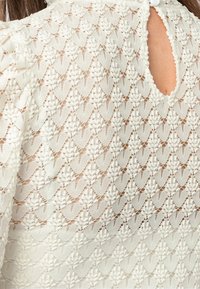 Vit spetsblus med ett texturerat mönster och scallopdesign. Detalj med nyckelhål bak med knappstängning. Lätt och delikat tyg.