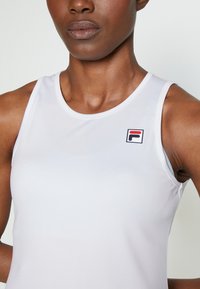 Hvit atletisk tanktopp med rund hals og ermeløs design. Vist logo i rødt og blått er plassert på den øvre venstre siden.
