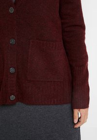 Bordowy kardigan wykonany z miękkiej dzianiny, z dekoltem w serek, zapięciem na guziki, ściągaczami na mankietach oraz przednią kieszenią naszywaną.