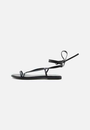 Sort flad sandal med tynde stropper, tå-loop og ankelsnøre på en ensartet lys baggrund.