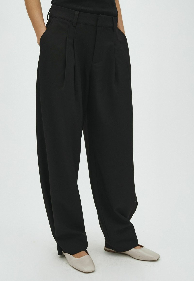 Personne portant un pantalon plissé noir large, les mains dans les poches et des chaussures plates beige clair sur un fond uni.