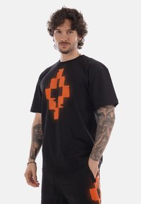 Hombre con cabello rizado y tatuajes que lleva una camiseta negra y pantalones cortos con un diseño brillante de cruz pixelada naranja, de pie con una mano en el bolsillo.