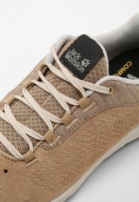 Tan fabric shoe with mesh and suede textures, featuring light laces, a patterned side, and a black logo label on the tongue.