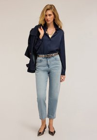 Donna bionda che indossa una blusa di raso blu navy, jeans a vita alta blu chiaro, collana d'oro e tacchi metallici a punta, mentre tiene un giubbotto scuro sulla spalla.