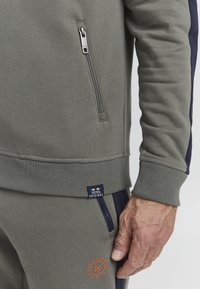 Gray sweatshirt with a zip pocket, ribbed cuffs, and logo label. Features dark blue side accents and orange embroidery on the pants.