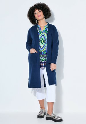 Frau mit lockigem Haar, die eine lange marineblaue Strickjacke über einer bunt gemusterten Bluse, weißen Cropped-Hosen und metallischen Plateau-Loafers trägt und steht.