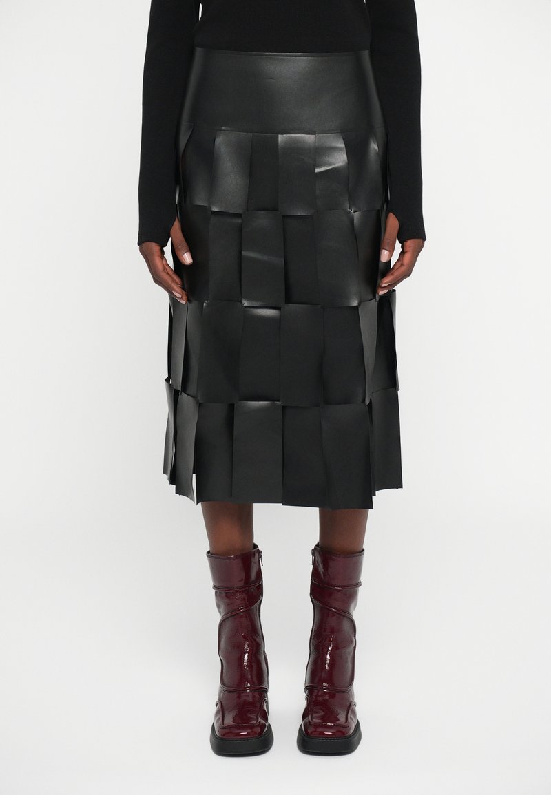 Falda midi negra con grandes paneles cuadrados cosidos, combinada con botas oscuras de cuero brillante hasta la mitad de la pantorrilla y una blusa negra de manga larga.