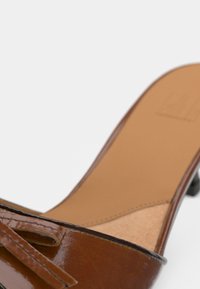 Καφέ δερμάτινο γυναικείο παπούτσι με τακούνι και γυαλιστερό φινίρισμα, με φαρδύ λουρί και μικρό φιόγκο στο μπροστινό μέρος. Λείο εσωτερικό επένδυμα.