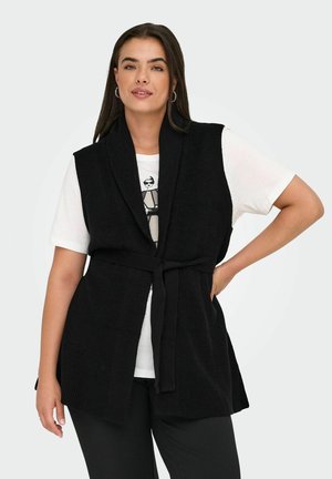 Femme portant un gilet en tricot sans manches noir noué à la taille sur un t-shirt graphique blanc et un pantalon noir, se tenant avec une main sur la hanche.