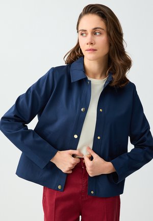 Kobieta ubrana w granatową zapinaną na guziki kurtkę na białą koszulę i czerwone spodnie, poprawiająca kurtkę, patrząc w bok na jednolitym tle.