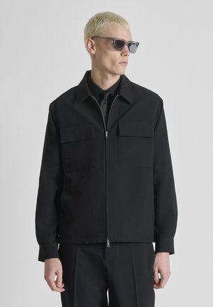 Blond person wearing black zip-up jacket with chest pockets, black shirt and tie, and dark sunglasses, looking to the side.